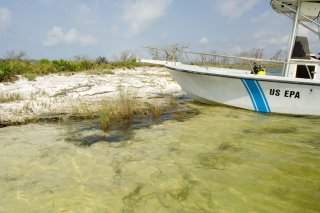 White boat with a blue strip on the side and U.S. EPA written next to it. The boat is pulled on on the shore of a beach with shite sand and grasses.