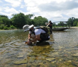 A man wearing a sunhat crouches down in a stream, a small boat and another person behind him
