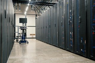 an aisle of server racks stands in a lit windowless space