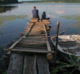 Image of an adult and a child sitting at the end of a dock fishing on a lake. 
