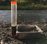 A stream with a rocky shore. There is a metal container with silt sitting on the shore with next to a test beakers with a water and sediment sample.